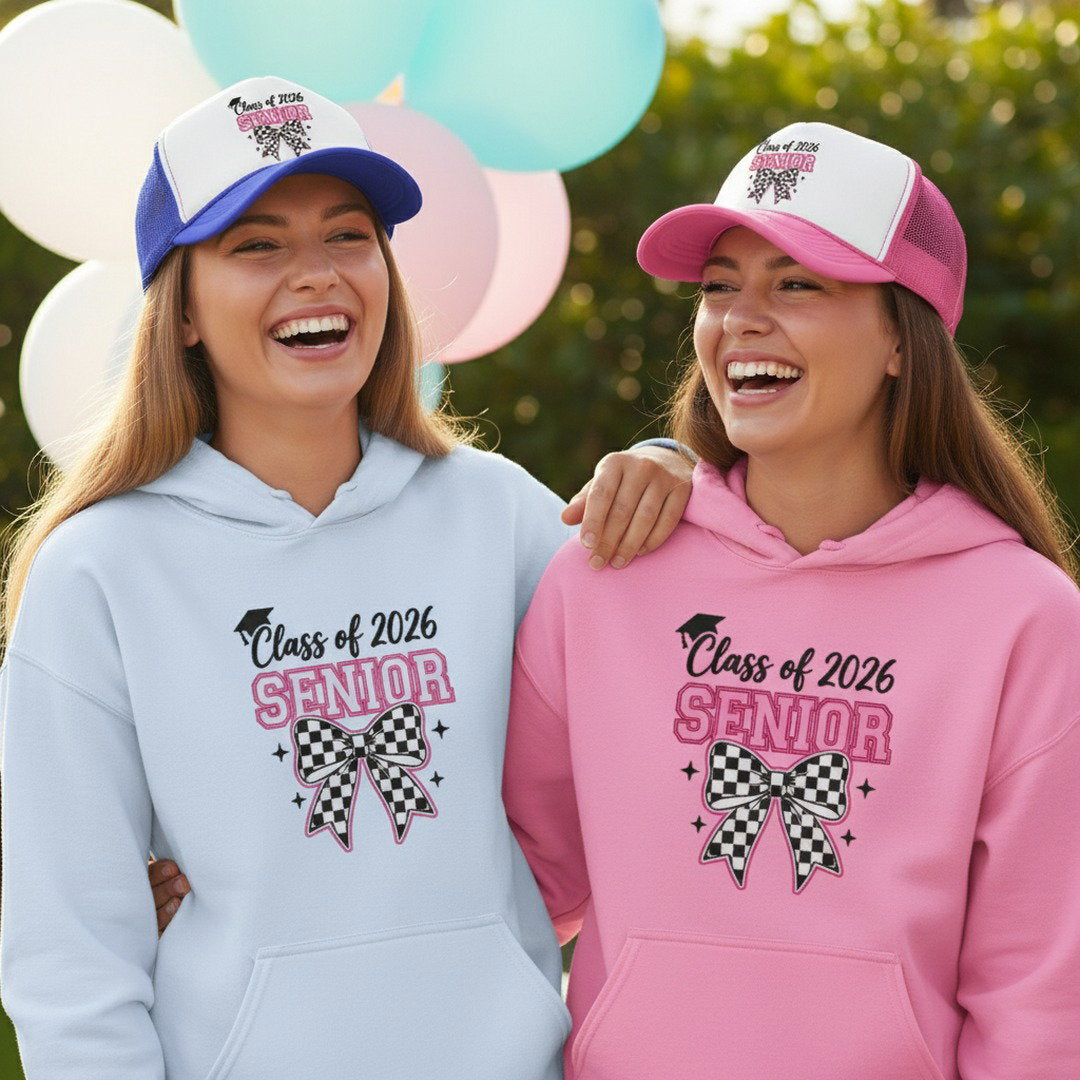 Two smiling young women wearing Class of 2026 Senior checkered bow hoodies in white and light pink with blue and pink Senior trucker caps laughing together at outdoor graduation party with colorful balloons and greenery in background