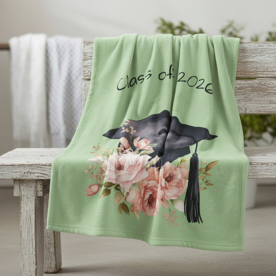 Class of 2026 sage green graduation blanket with pink floral cap design on rustic wooden bench, outdoor lifestyle photo
