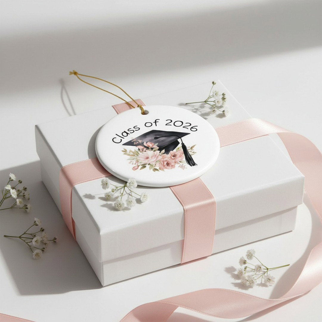 Class of 2026 graduation ceramic ornament resting on white gift box with pink satin ribbon and baby's breath flowers, elegant graduation gift presentation