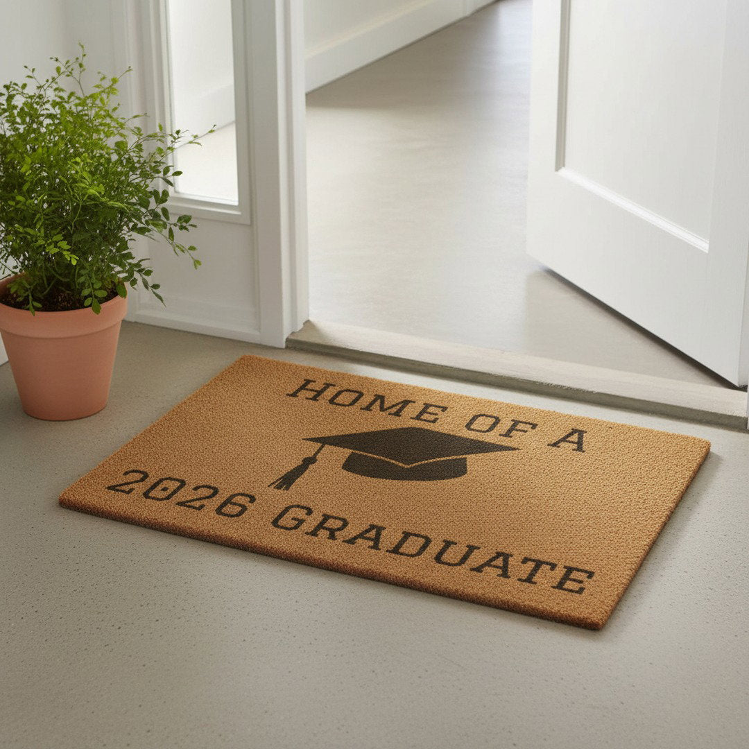 Home of a 2026 Graduate coir doormat at white front door beside terracotta potted plant, natural fiber graduation welcome mat