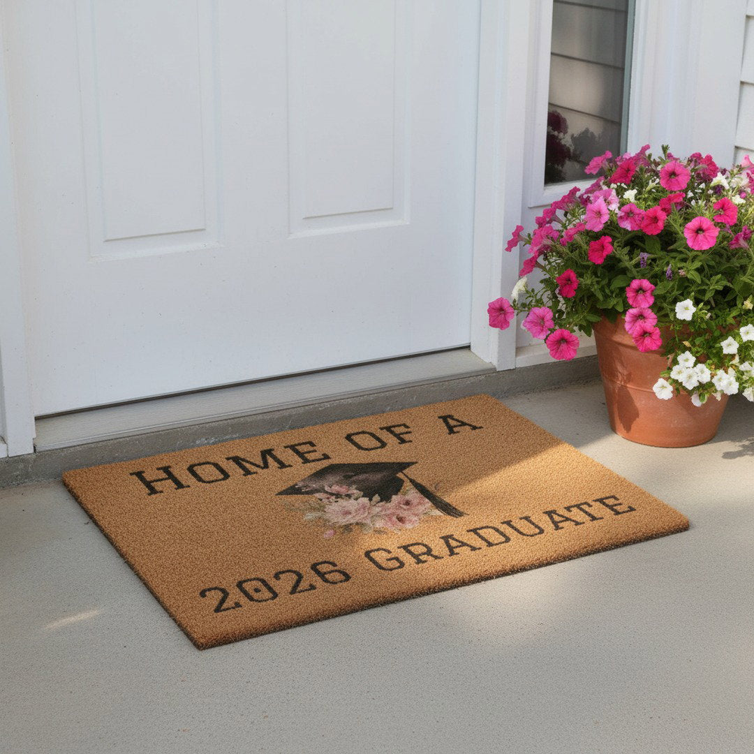 Home of a 2026 Graduate floral doormat at white front door beside terracotta pot overflowing with pink petunias, graduation home decor