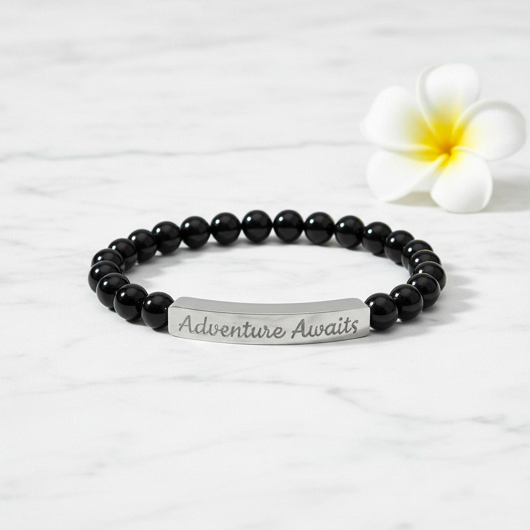 Engraved Natural Stone Stretch Bracelet with glossy black onyx beads and a polished silver stainless steel bar engraved with "Adventure Awaits" in script, flat lay on white marble with a small white flower accent