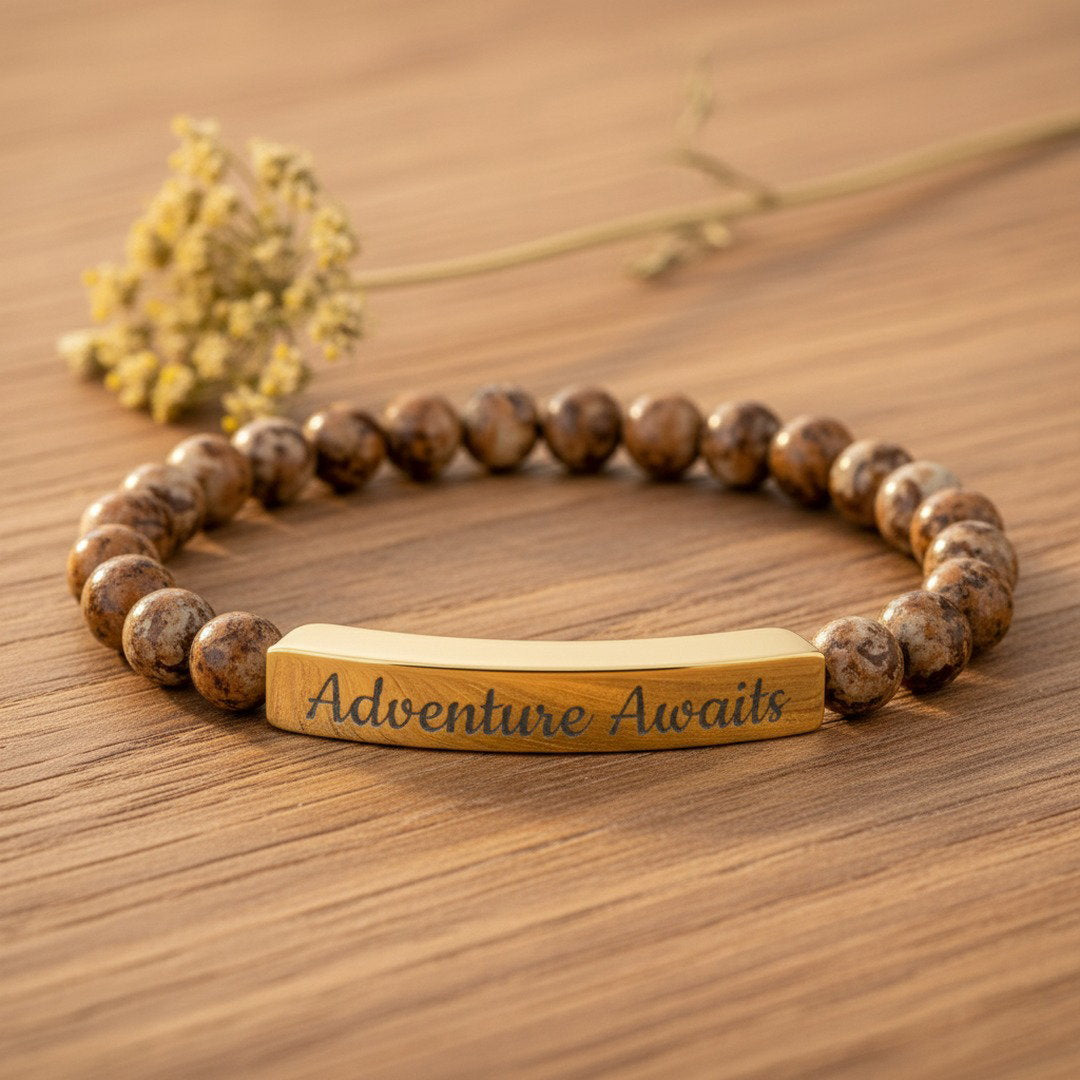 Engraved Natural Stone Stretch Bracelet with warm brown picture jasper beads and an 18K gold PVD bar engraved with "Adventure Awaits" in script, flat lay on natural wood with a dried wildflower accent