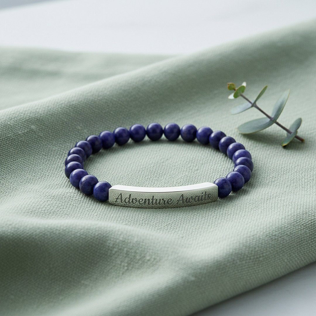 Engraved Natural Stone Stretch Bracelet with deep blue sodalite beads and a polished silver stainless steel bar engraved with "Adventure Awaits" in script, displayed on sage green linen with a eucalyptus sprig