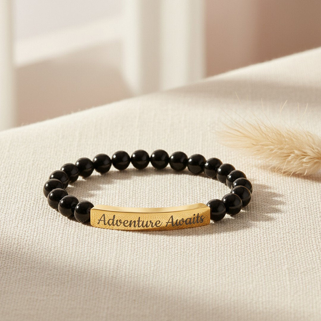 Engraved Natural Stone Stretch Bracelet with glossy black onyx beads and an 18K gold PVD bar engraved with "Adventure Awaits" in script, displayed on soft ivory linen with a dried pampas grass stem, warm natural light