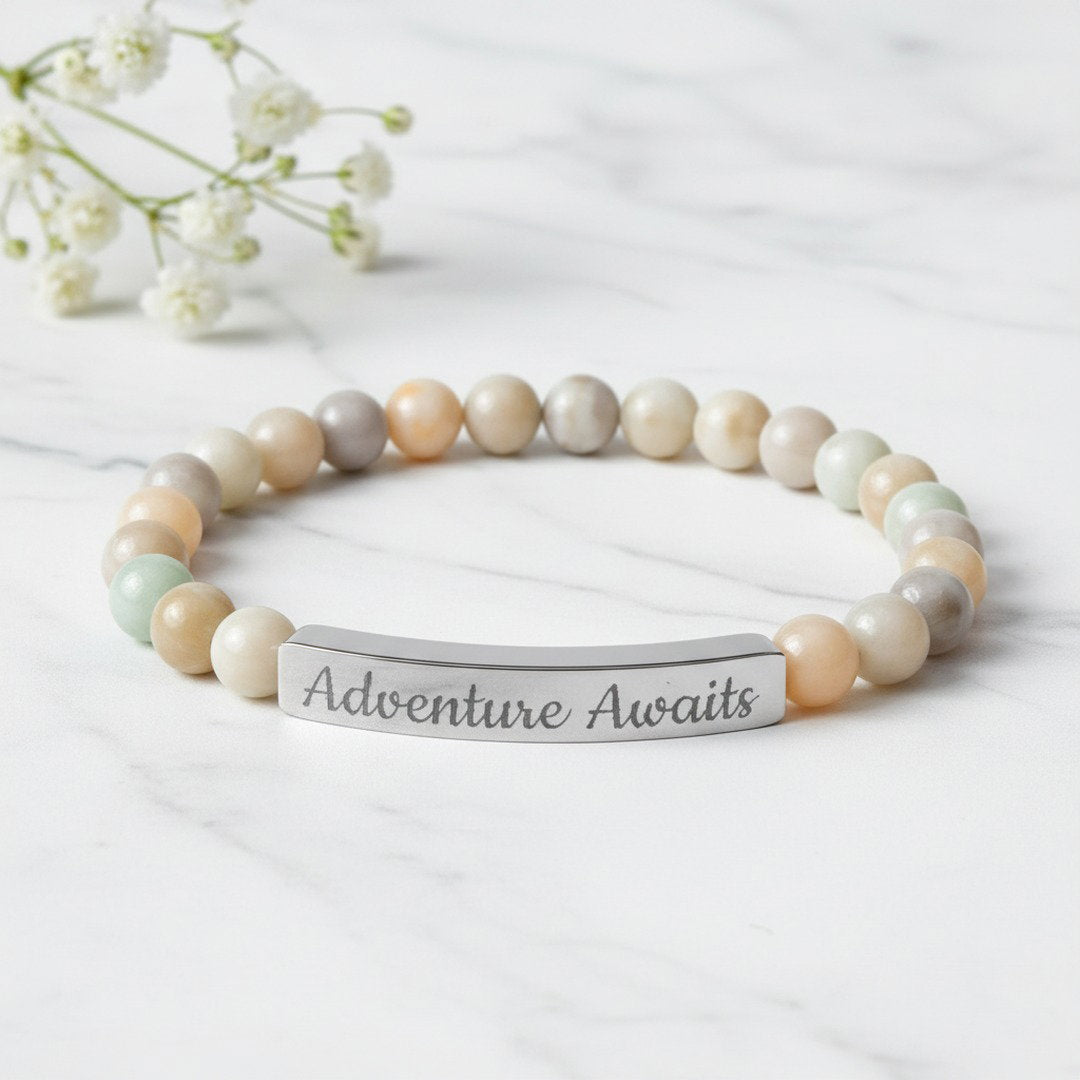 Engraved Natural Stone Stretch Bracelet with soft multicolor amazonite beads in ivory, peach, and sage tones and an 18K gold PVD bar engraved with "Adventure Awaits" in script, styled on warm linen with a white ranunculus bloom