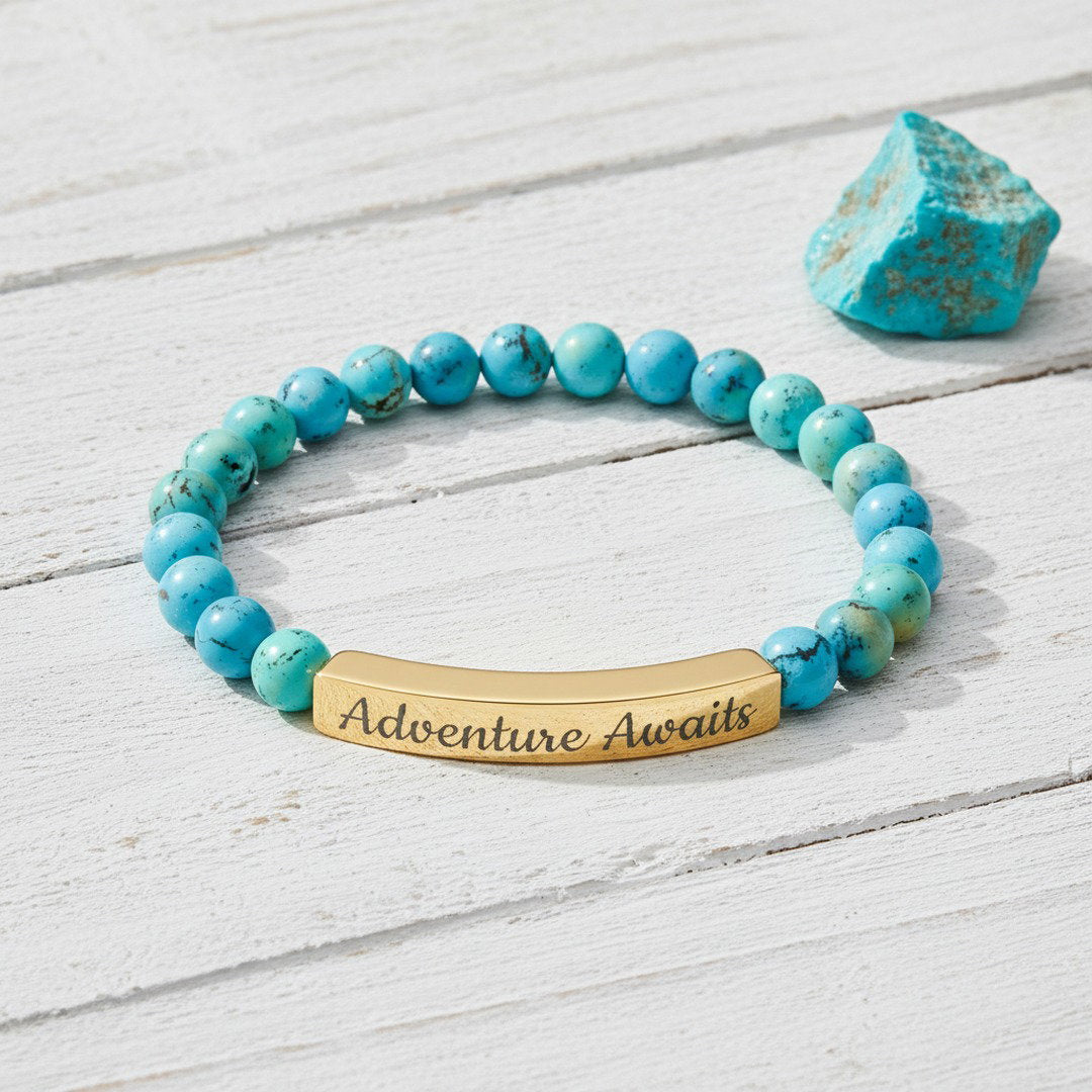 Engraved Natural Stone Stretch Bracelet with vibrant blue turquoise beads and an 18K gold PVD bar engraved with "Adventure Awaits" in script, flat lay on white wood with a raw turquoise crystal accent