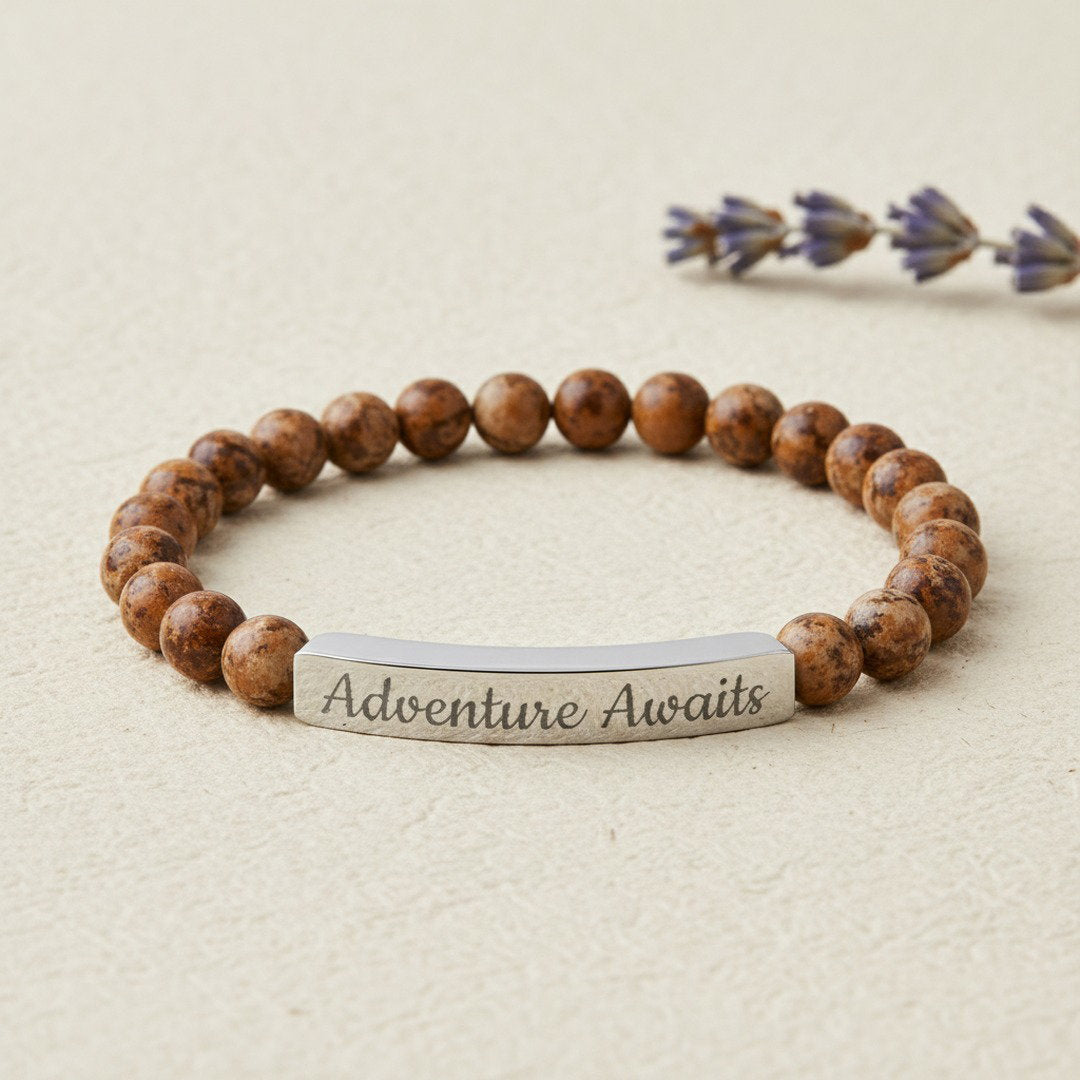 Engraved Natural Stone Stretch Bracelet with warm brown picture jasper beads and a polished silver stainless steel bar engraved with "Adventure Awaits" in script, styled on a cream textured surface with dried lavender