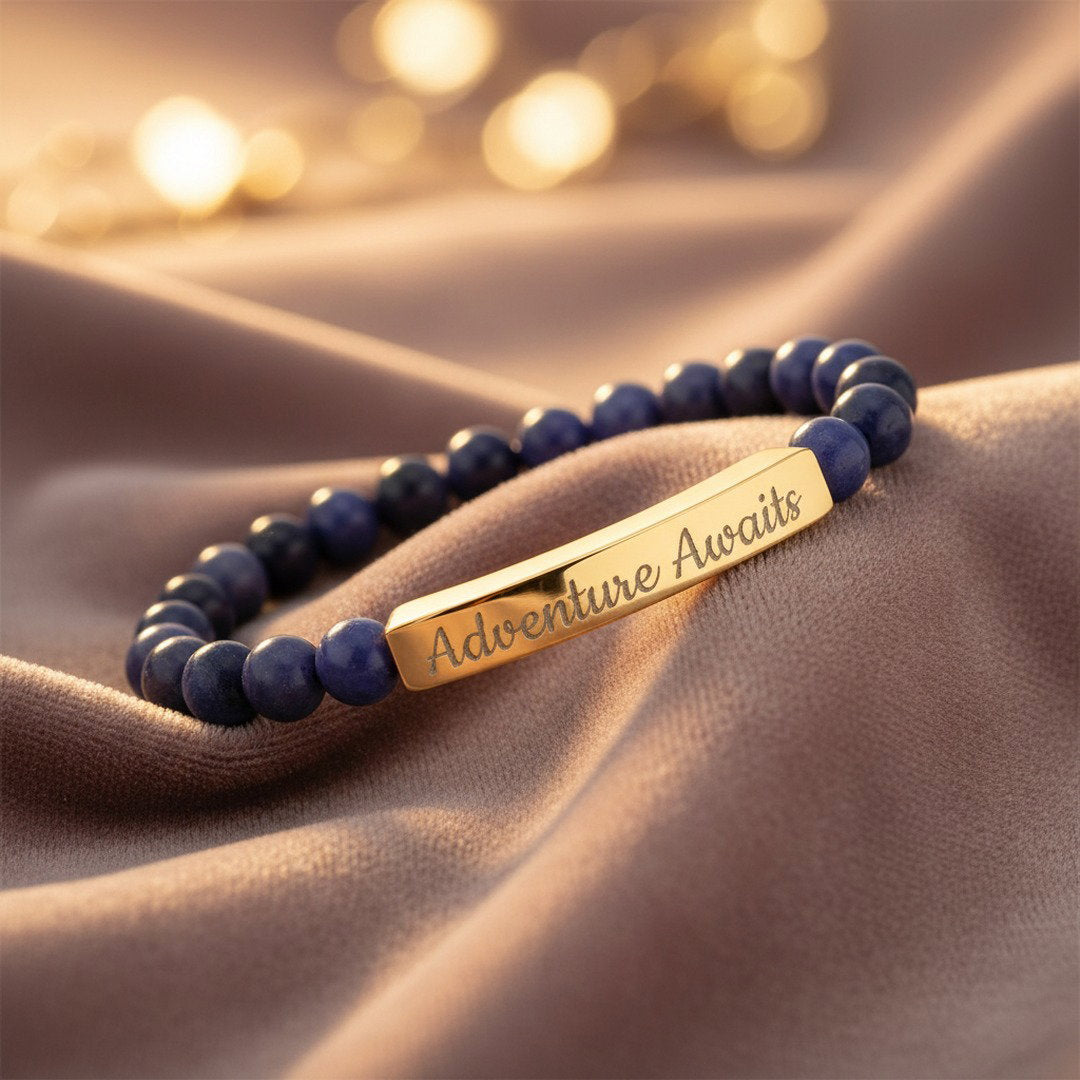 Engraved Natural Stone Stretch Bracelet with deep blue and purple sodalite beads and an 18K gold PVD bar engraved with "Adventure Awaits" in script, styled on dusty mauve velvet with soft warm golden light, editorial jewelry photography