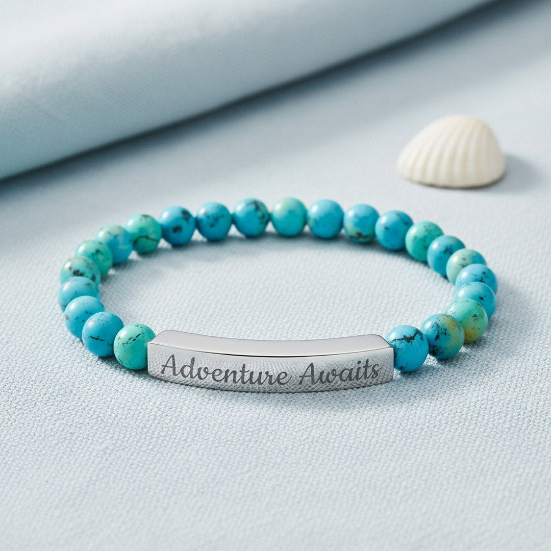 Engraved Natural Stone Stretch Bracelet with vibrant blue turquoise beads and a polished silver stainless steel bar engraved with "Adventure Awaits" in script, displayed on cool blue linen with a white shell accent