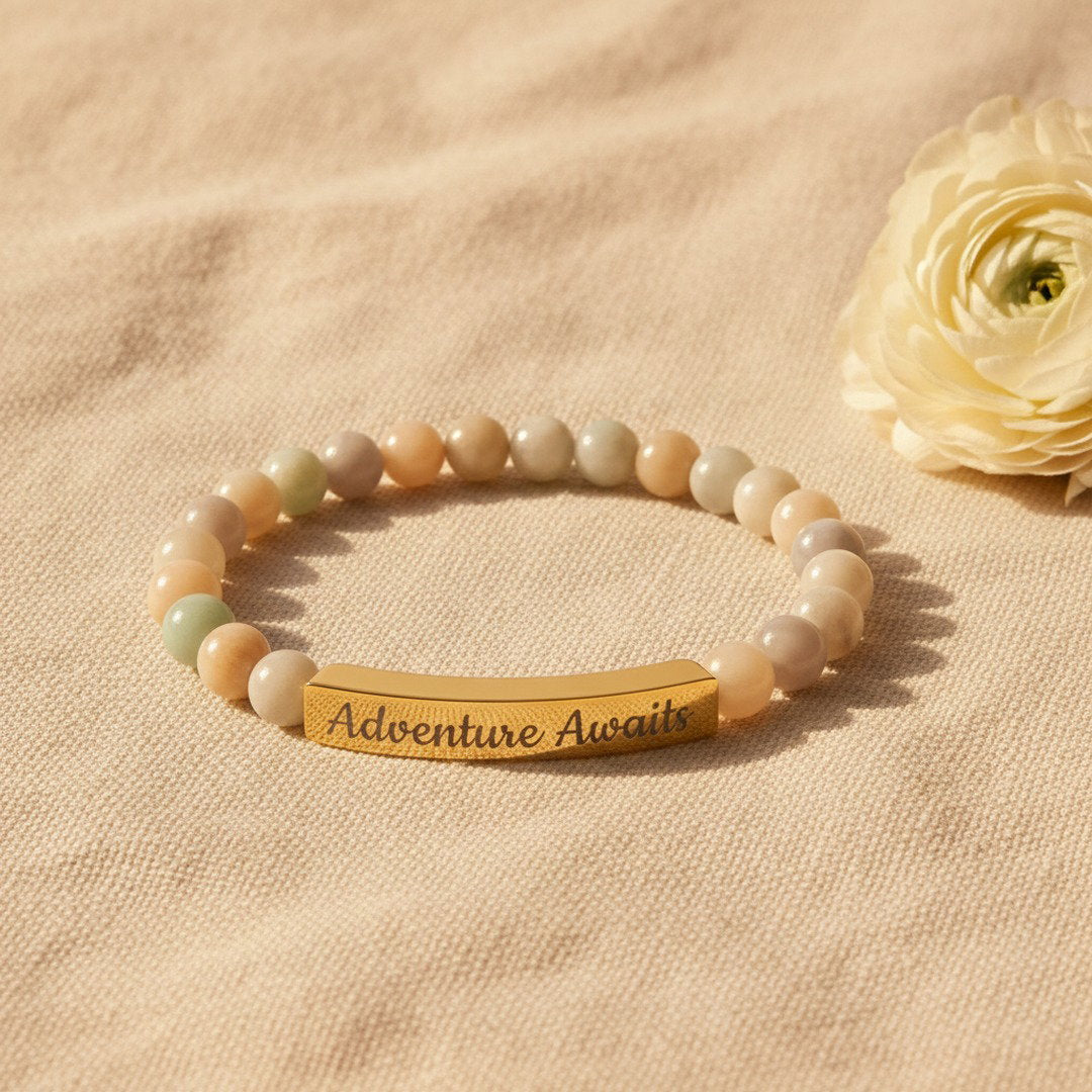 Engraved Natural Stone Stretch Bracelet with soft multicolor amazonite beads in creamy ivory, pale peach, and sage green tones and an 18K gold PVD bar engraved with "Adventure Awaits" in script, styled on warm blush linen with a white ranunculus bloom, soft golden natural light