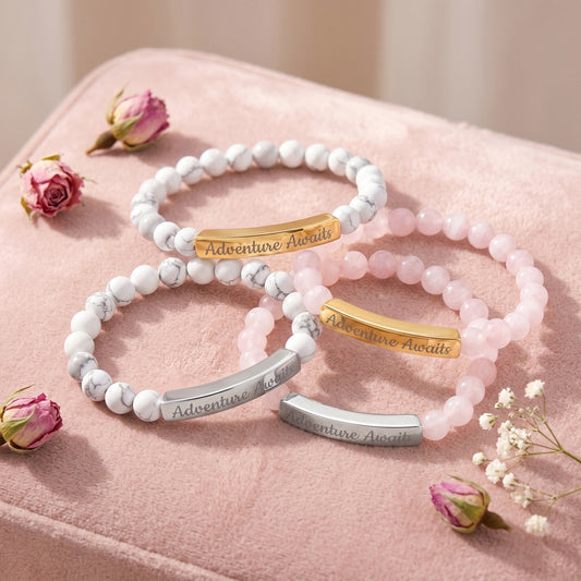 Engraved "Adventure Awaits" Natural Stone Stretch Bracelets in four variants — white howlite with gold bar, white howlite with silver bar, pink rose quartz with gold bar, and pink rose quartz with silver bar — arranged in an organic cluster on blush pink velvet with scattered dried rosebuds and baby's breath, soft natural light, editorial jewelry photography