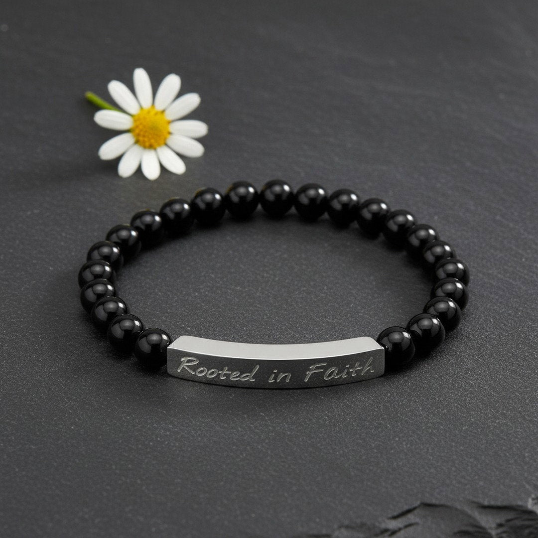 A stretch bracelet with glossy black onyx beads and a polished silver stainless steel bar engraved with "Rooted in Faith" in script, flat lay on a dark slate surface with a single small white flower accent, moody soft natural light, editorial jewelry photography, bold and faith-inspired aesthetic