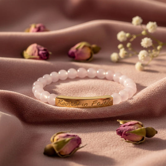 A stretch bracelet with soft blush pink rose quartz beads with a gentle translucent glow and a curved 18K gold bar engraved with "Rooted in Faith" in elegant script, styled on a dusty rose velvet surface with scattered dried pink rosebuds and a sprig of baby's breath, warm soft natural light, romantic editorial jewelry photography, feminine and faith-inspired