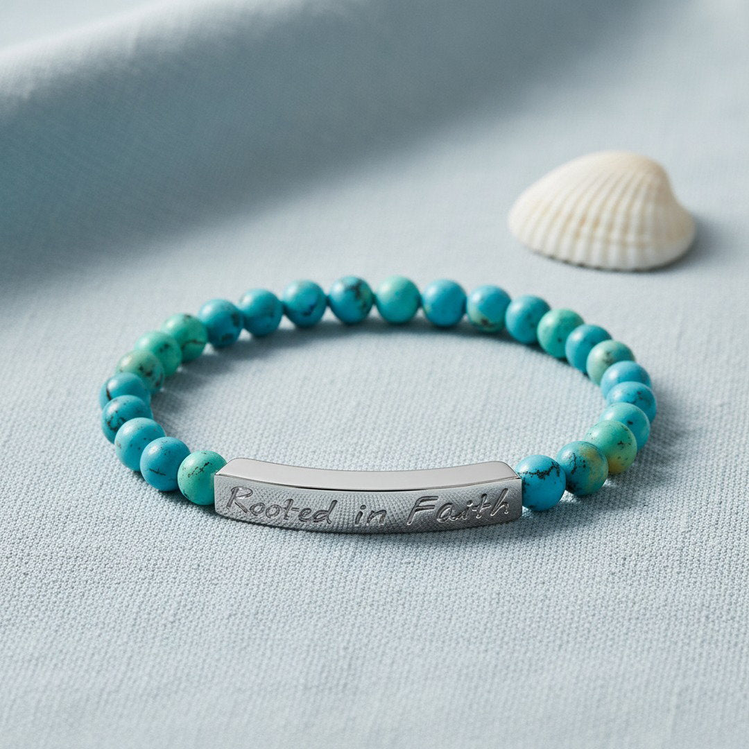 Rooted in Faith engraved natural stone stretch bracelet with vibrant blue turquoise beads featuring natural dark matrix veining and a polished silver stainless steel bar, displayed on a cool blue-gray linen surface with a white seashell accent, soft diffused natural light, clean coastal editorial jewelry photography