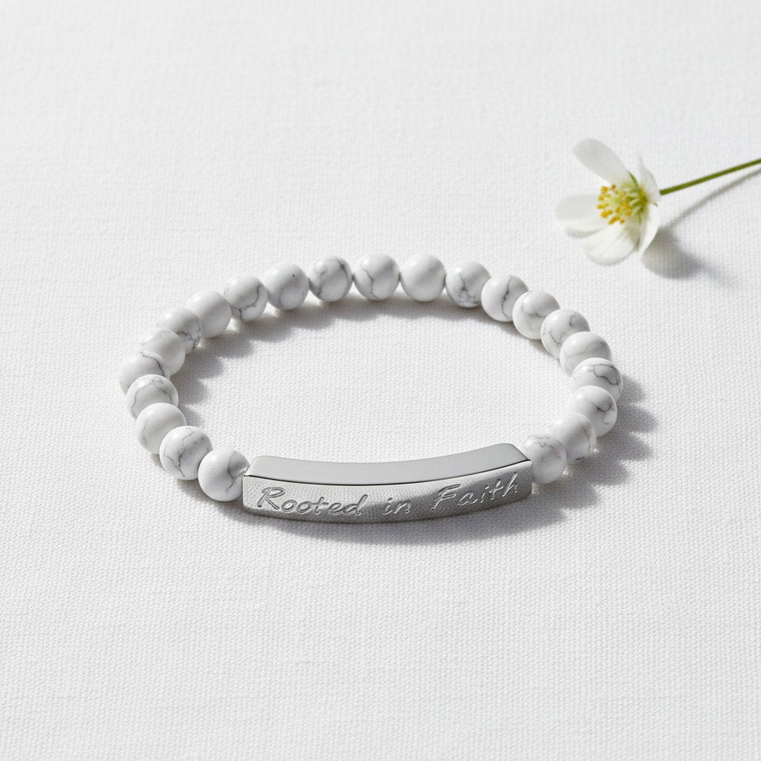 Rooted in Faith engraved natural stone stretch bracelet with smooth white howlite beads featuring subtle gray veining and an 18K gold bar, flat lay on warm cream linen with a white ranunculus bloom, soft golden natural light, premium editorial jewelry photography