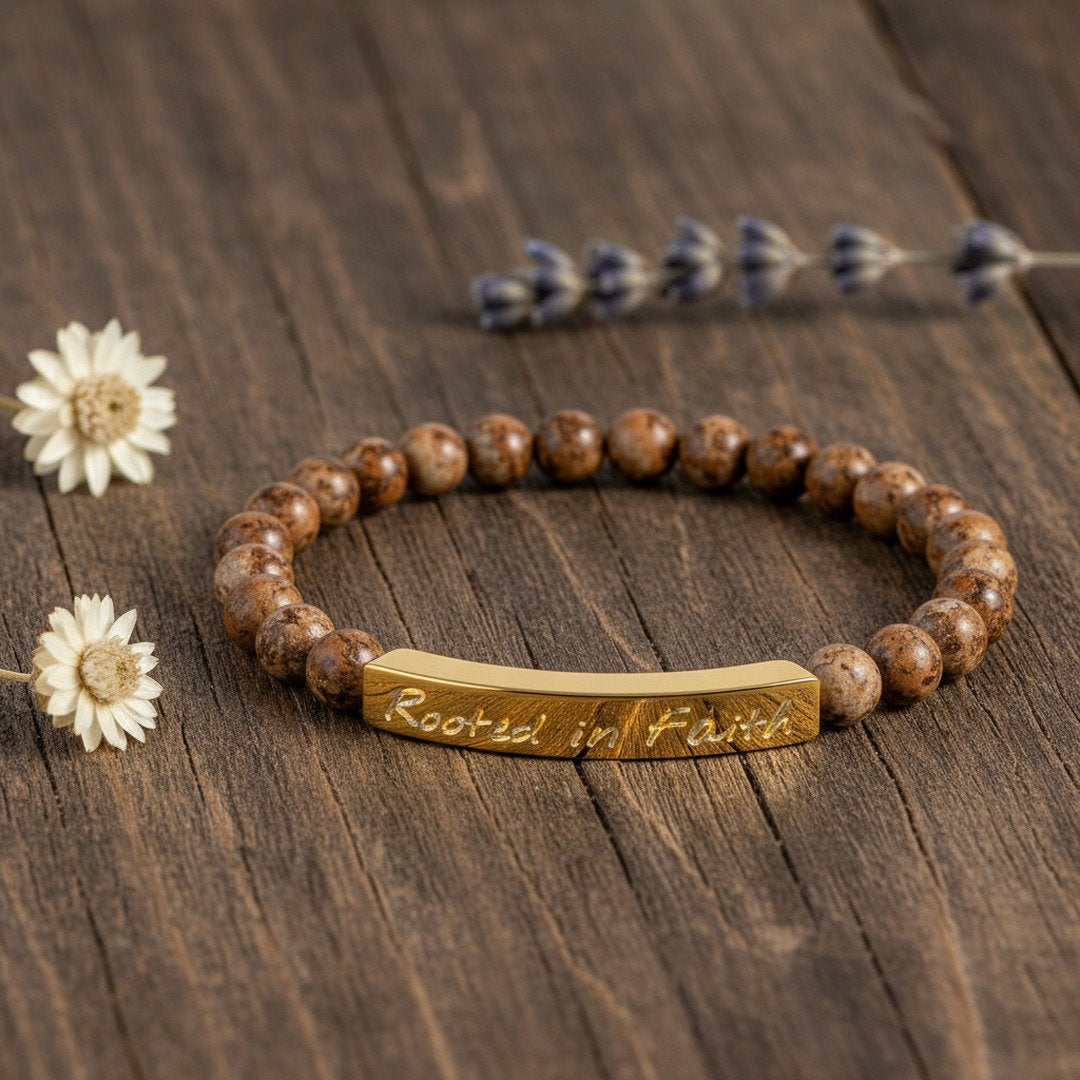 Rooted in Faith engraved natural stone stretch bracelet with warm brown picture jasper beads and an 18K gold bar, flat lay on a dark wood grain surface with dried white daisies and a lavender sprig, warm earthy tones, premium editorial jewelry photography