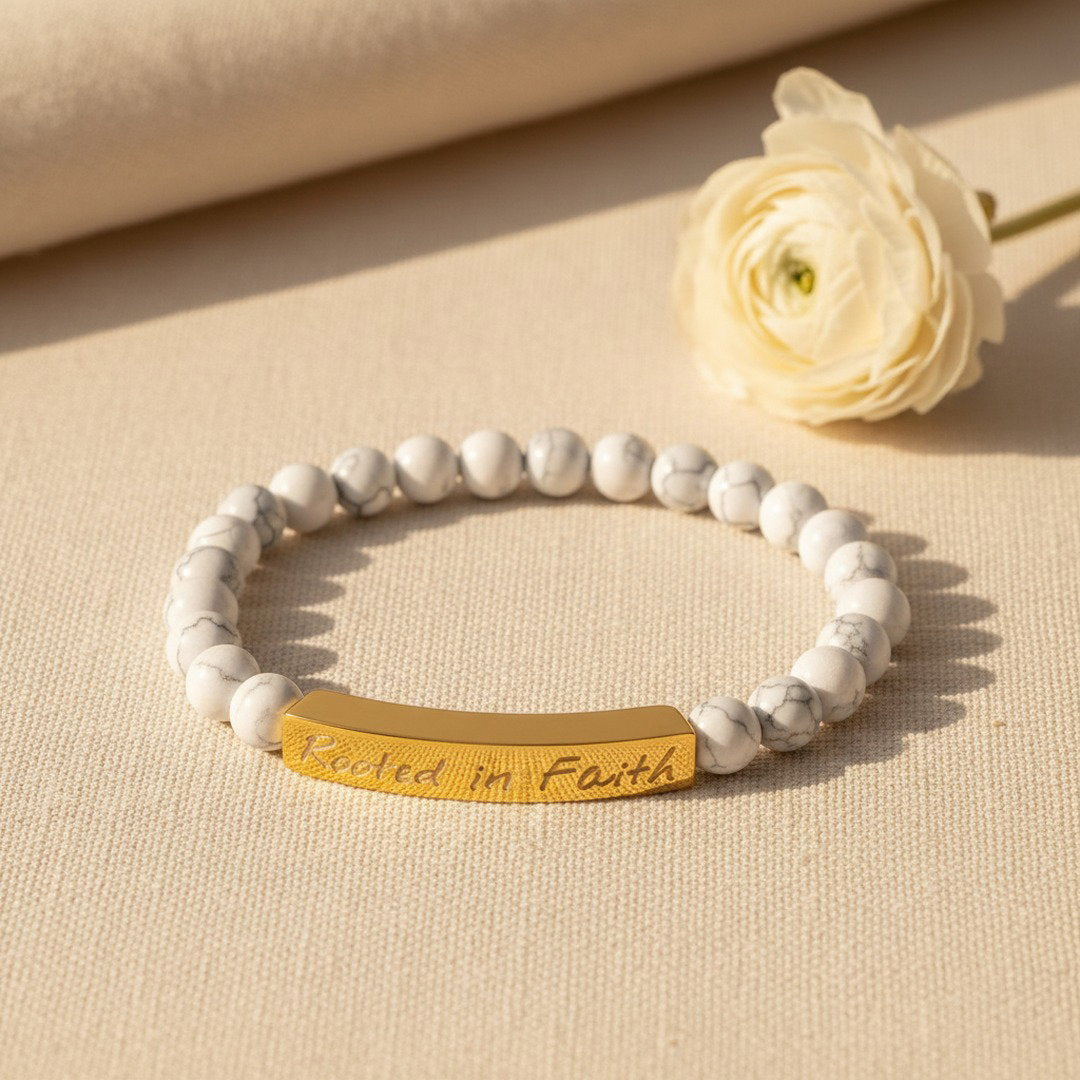 A stretch bracelet with smooth white howlite beads featuring subtle gray veining and a curved 18K gold bar engraved with "Rooted in Faith" in elegant script, flat lay on a warm cream linen surface with a small dried white ranunculus bloom, soft golden natural light, premium editorial jewelry photography, pure and feminine