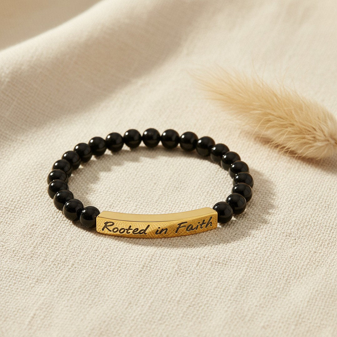 A stretch bracelet with glossy black onyx beads and a curved 18K gold bar engraved with "Rooted in Faith" in elegant script, displayed on a soft ivory linen surface with a dried pampas grass stem, warm natural light, bold and faith-inspired editorial jewelry photography