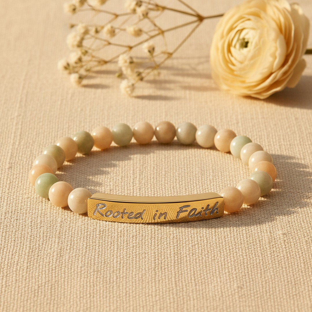Rooted in Faith engraved natural stone stretch bracelet with soft multicolor amazonite beads and an 18K gold bar, styled on warm ivory linen with a white ranunculus bloom, soft golden natural light, premium editorial jewelry photography