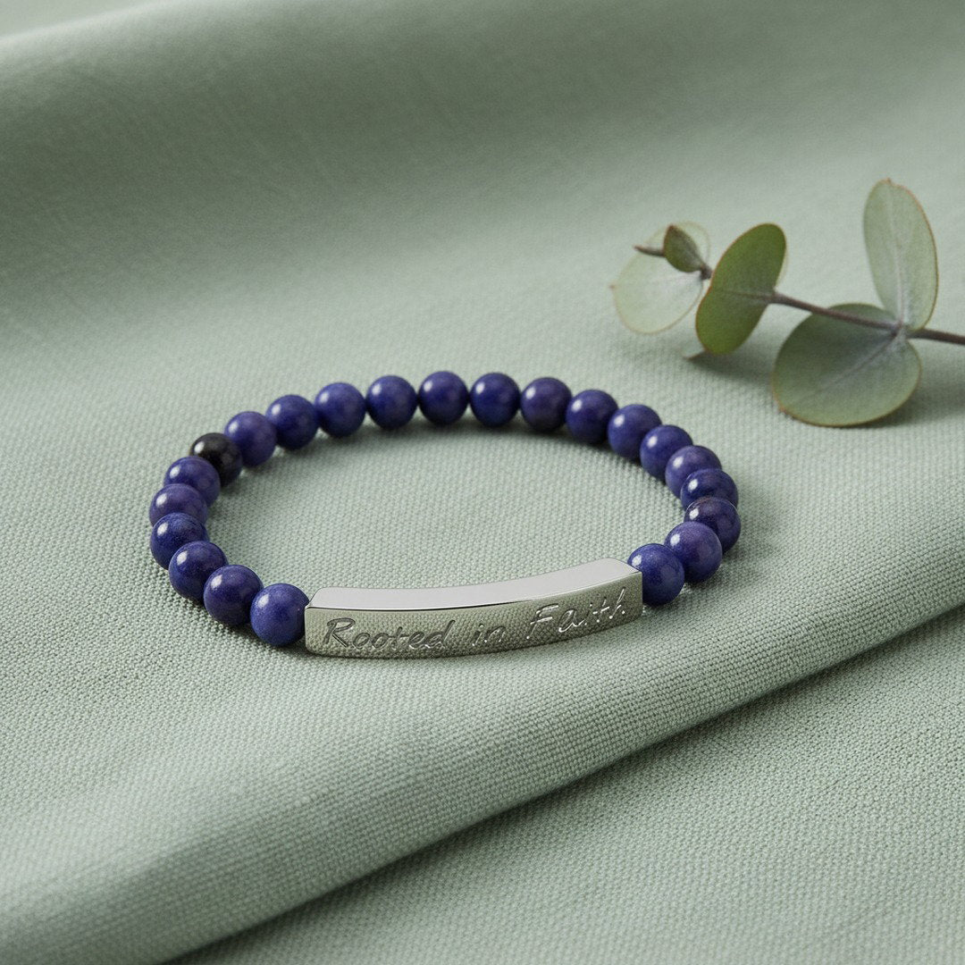 Rooted in Faith engraved natural stone stretch bracelet with deep blue and purple sodalite beads and a polished silver stainless steel bar, displayed on a sage green linen cloth with a fresh eucalyptus sprig, cool diffused natural light, clean editorial jewelry photography