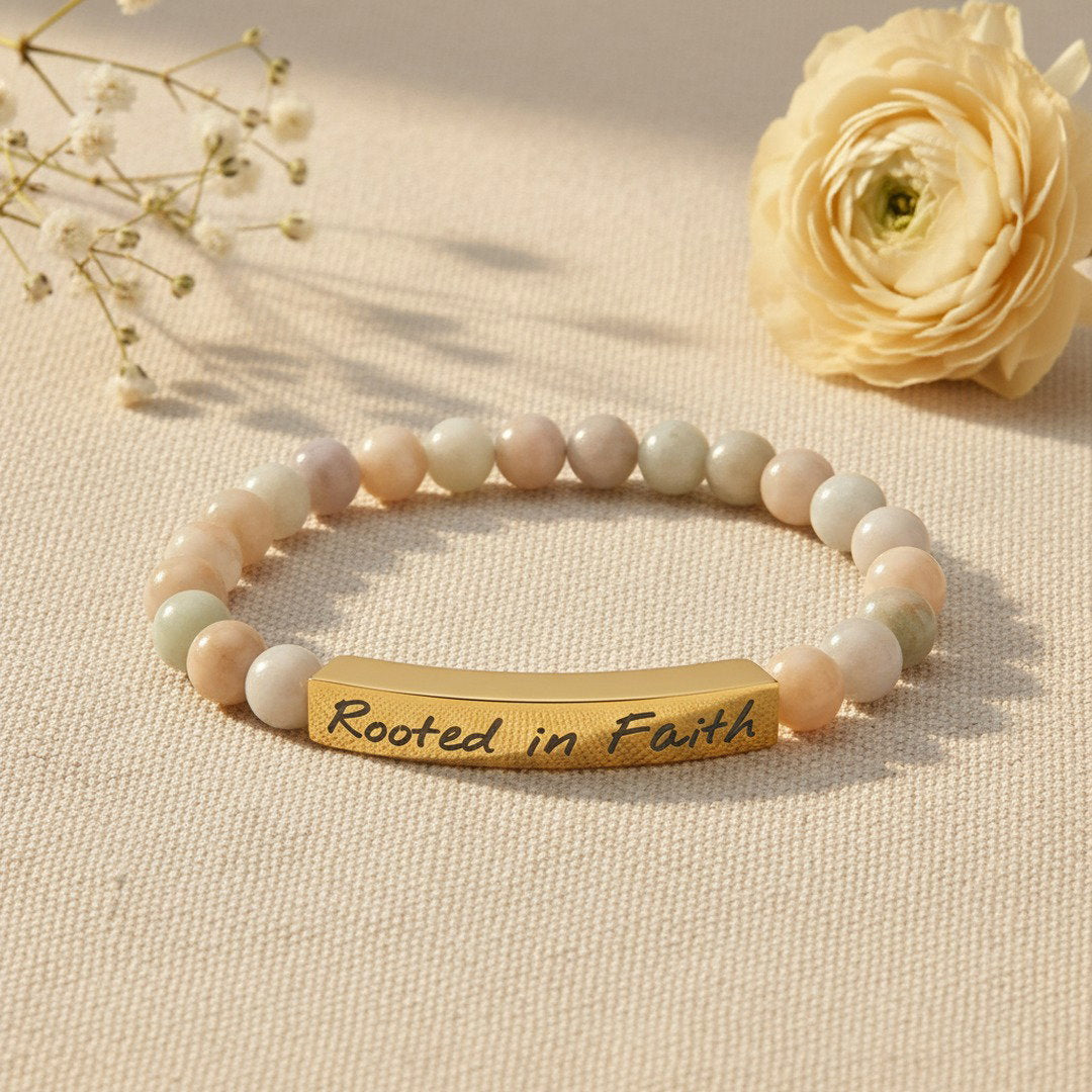 Rooted in Faith engraved natural stone stretch bracelet with soft multicolor amazonite beads in creamy ivory, pale peach, and sage green tones and an 18K gold PVD bar engraved with "Rooted in Faith" in elegant script, styled on warm ivory linen with a dried white ranunculus bloom, soft golden natural light, premium editorial jewelry photography, delicate and feminine