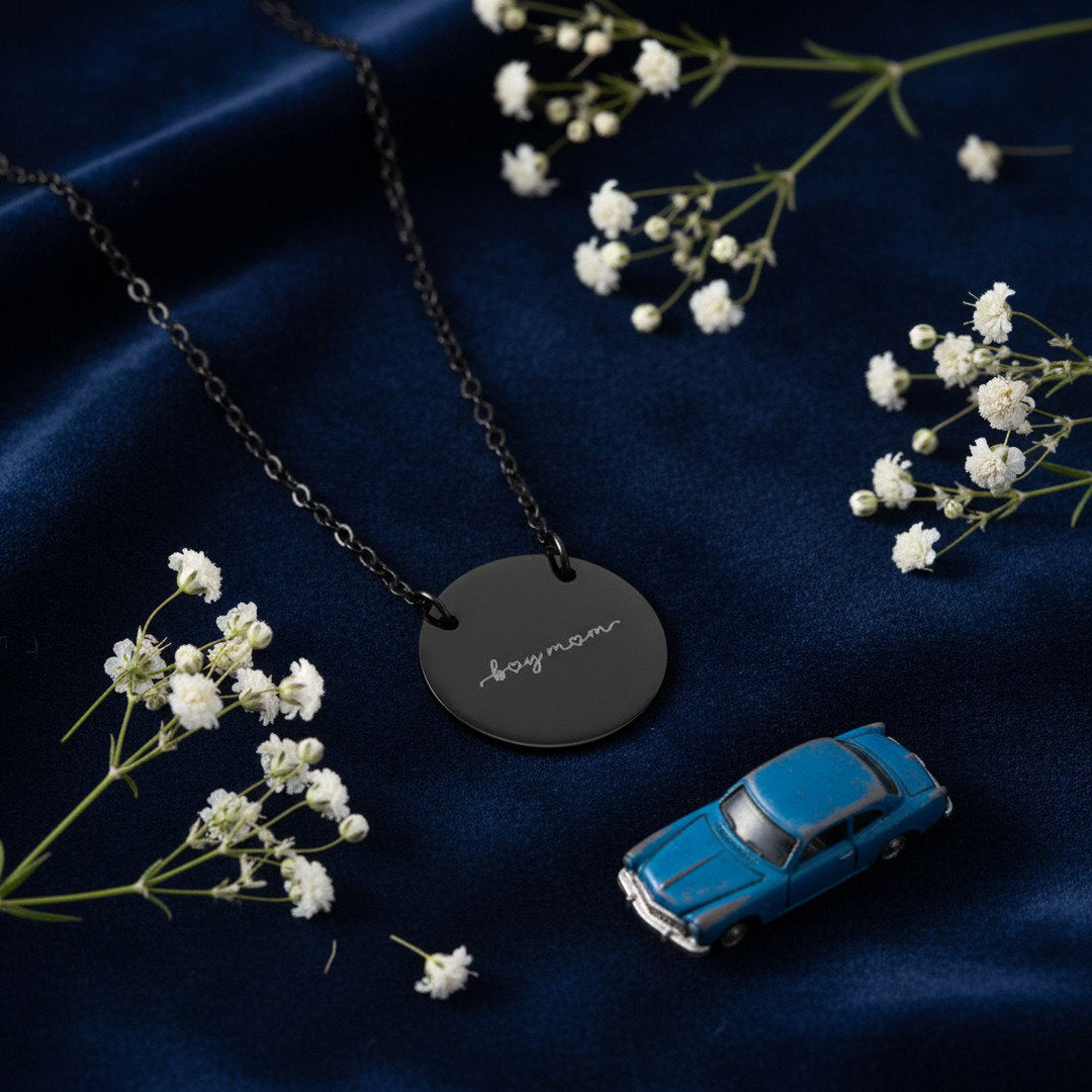 Engraved "Boy Mom" disc necklace in black, flat lay on navy surface with white wildflowers, gift for boy moms