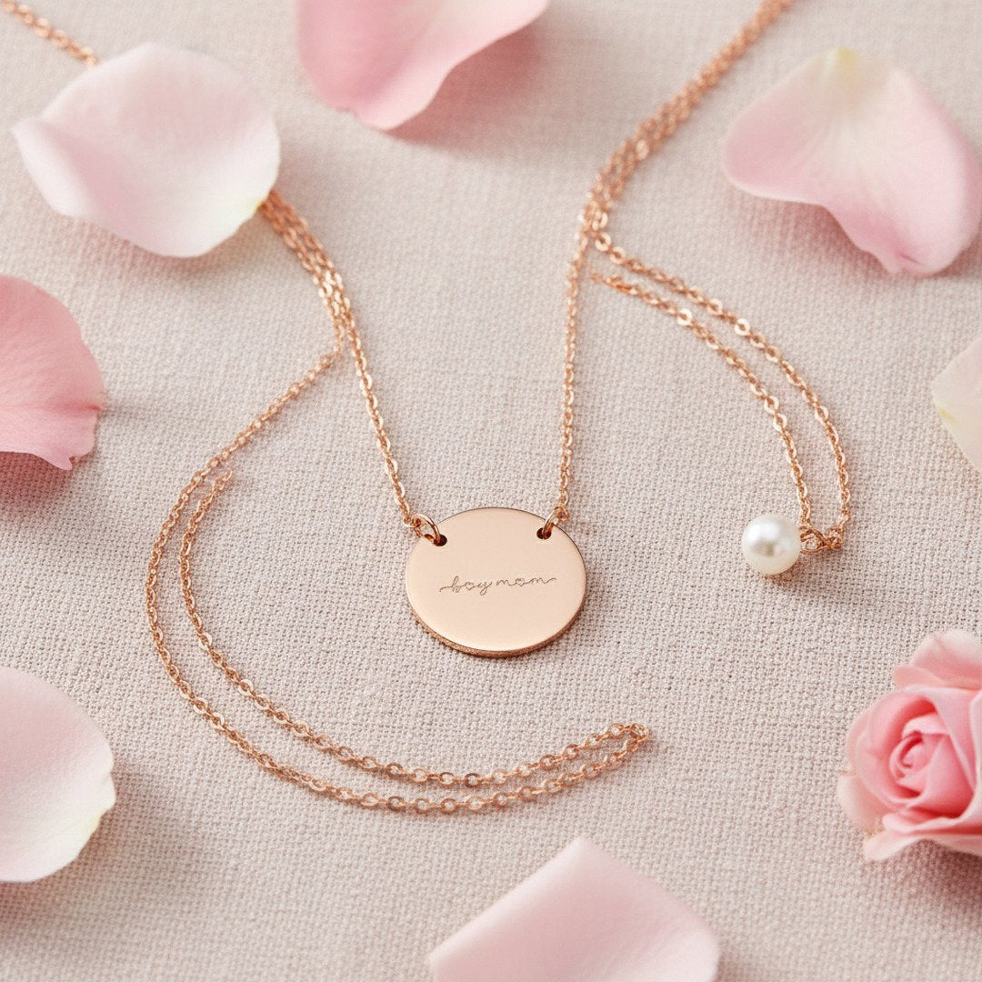 Engraved "Boy Mom" disc necklace in rose gold, flat lay on blush linen with pink rose petals and pearl accent, romantic Mother's Day jewelry gift