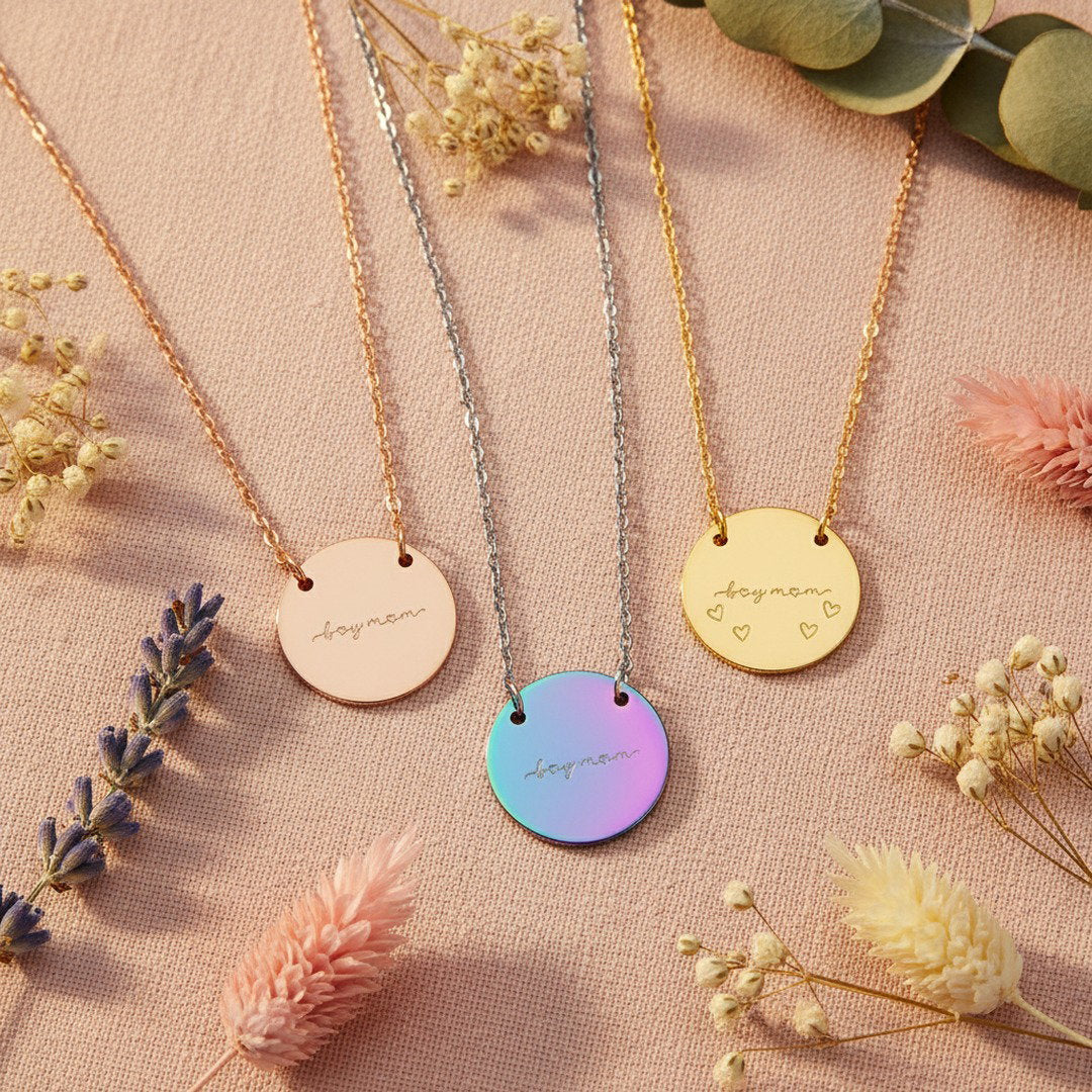Three "Boy Mom" disc necklaces in rose gold, rainbow, and gold arranged on blush linen with dried wildflowers and eucalyptus, Mother's Day jewelry gift set