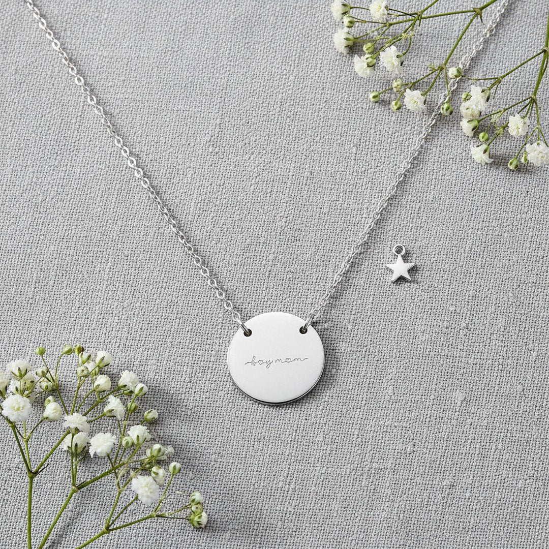 Engraved "Boy Mom" disc necklace in silver with star charm, flat lay on gray linen with baby's breath, gift for boy moms