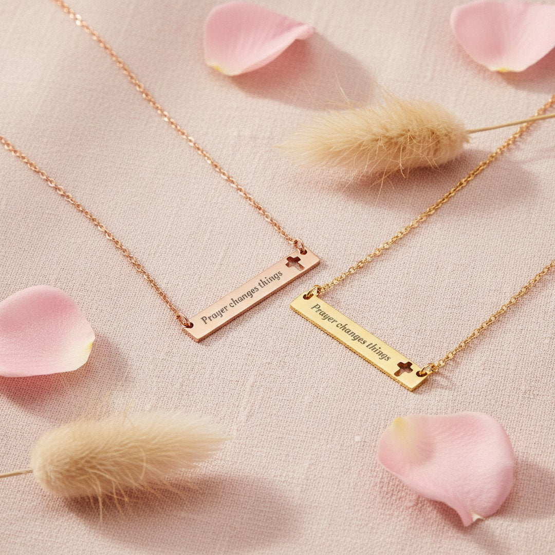 Two "Prayer Changes Things" bar necklaces in rose gold and gold with cut-out cross, styled flat lay on blush linen with pink rose petals and pampas grass, faith jewelry gift set