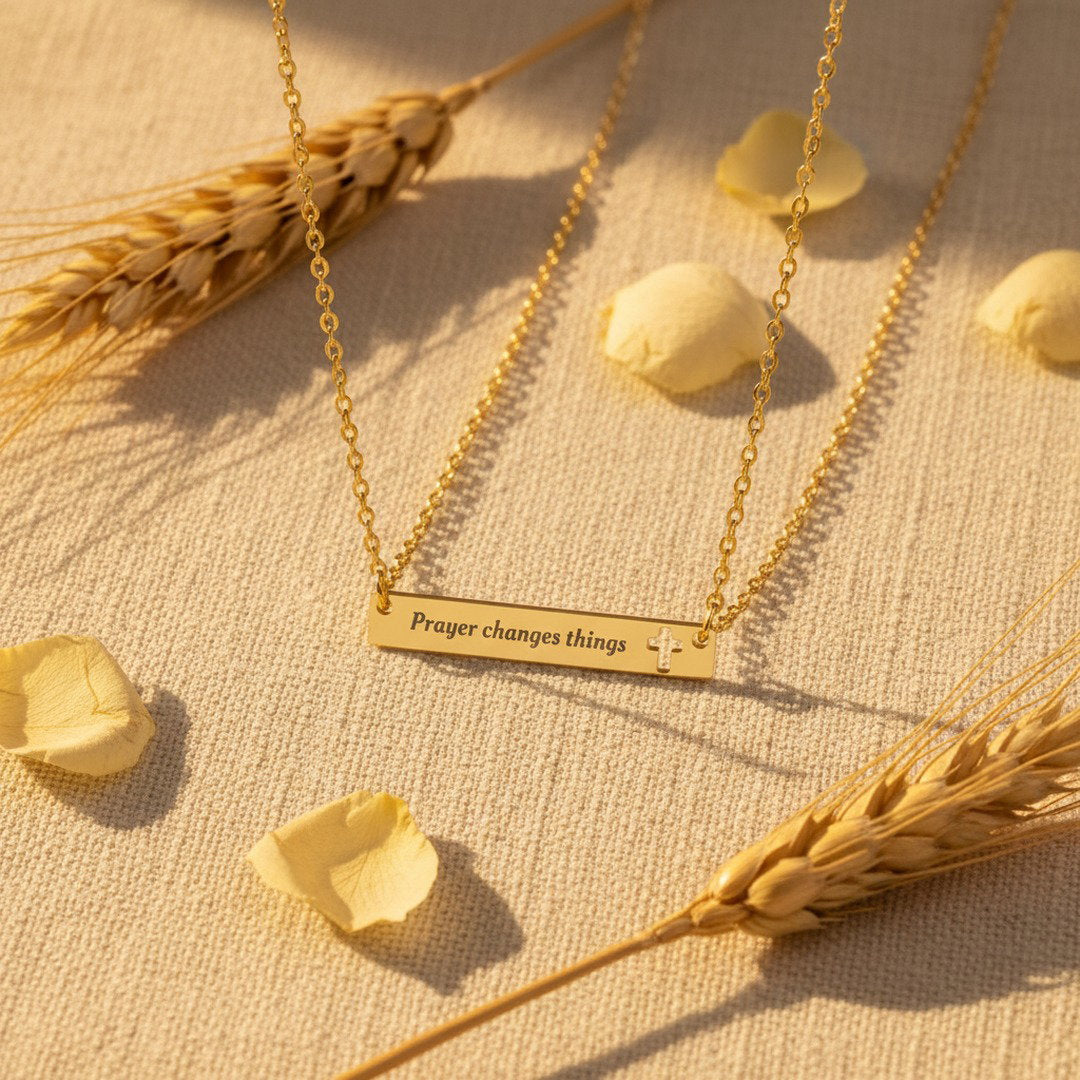 Engraved "Prayer Changes Things" bar necklace in gold with cut-out cross, flat lay on blush linen with pink rose petals and pampas grass, faith jewelry gift