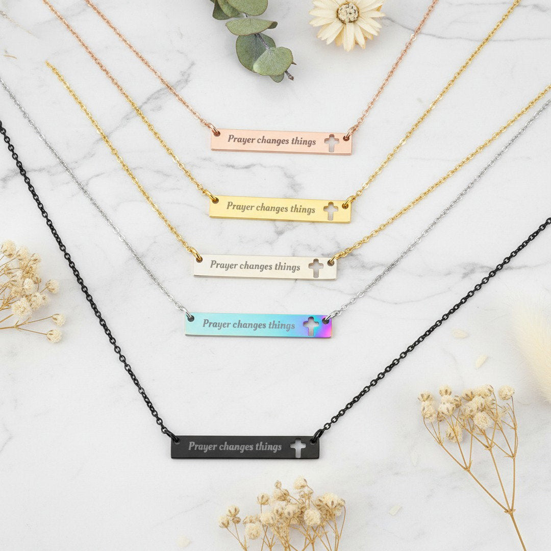 All five "Prayer Changes Things" bar necklaces in rose gold, gold, silver, rainbow, and black arranged in a fan on white marble with dried florals, complete faith necklace collection