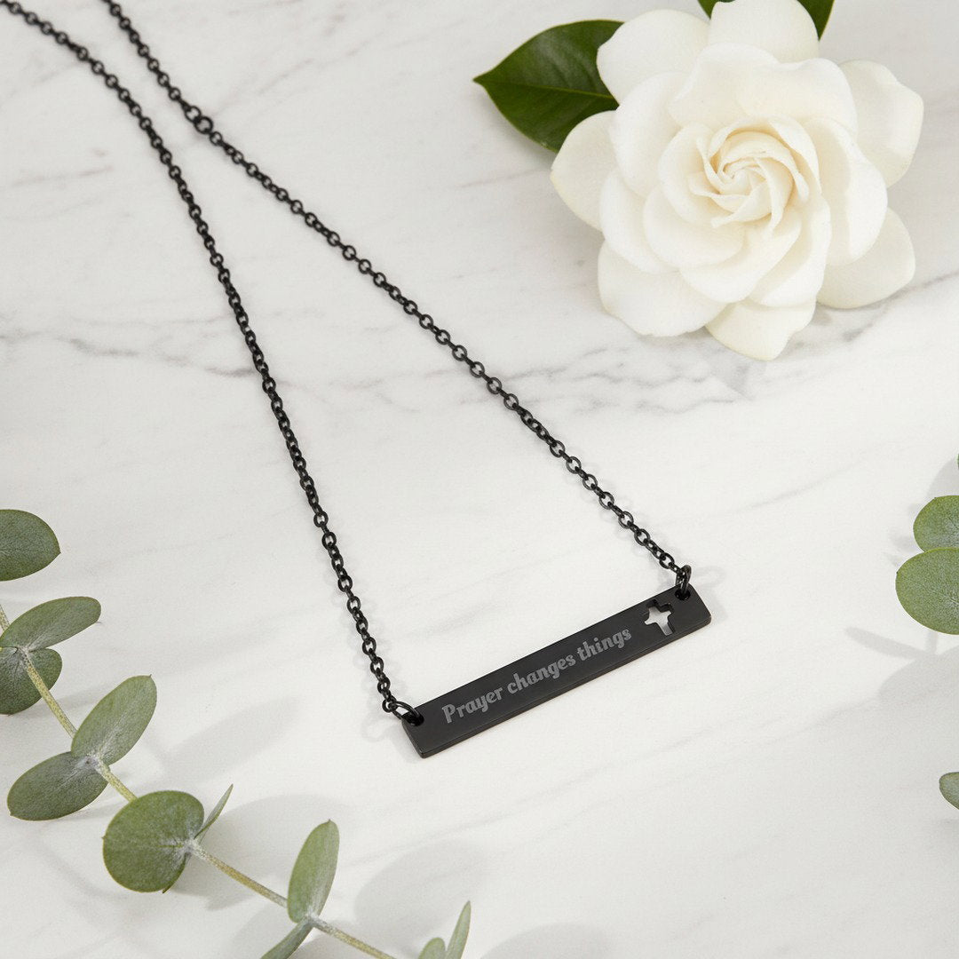Engraved "Prayer Changes Things" bar necklace in black with cut-out cross, flat lay on white marble with white wildflowers, faith jewelry gift

