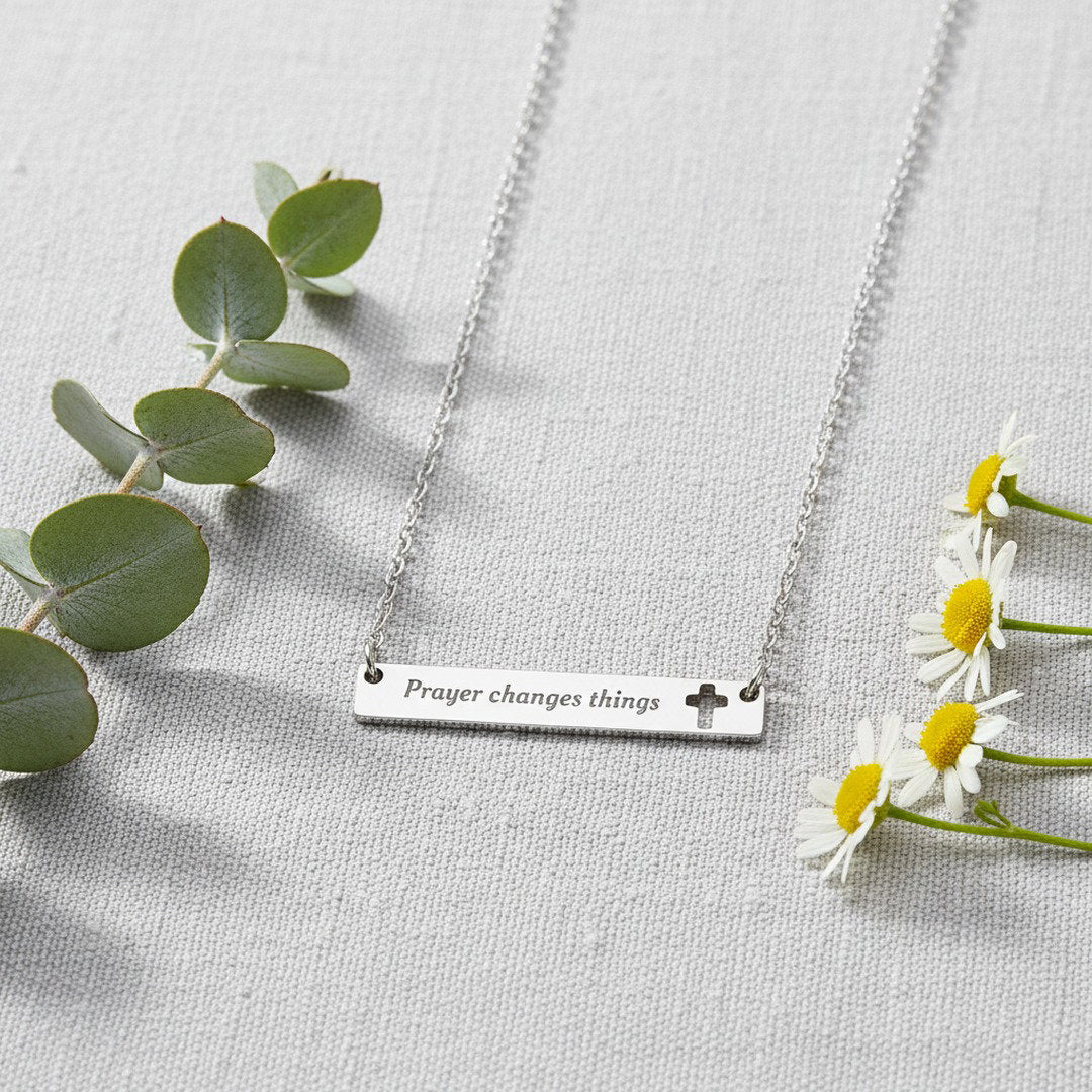 Engraved "Prayer Changes Things" bar necklace in silver with cut-out cross, flat lay on gray linen with eucalyptus and white daisies, faith jewelry gift