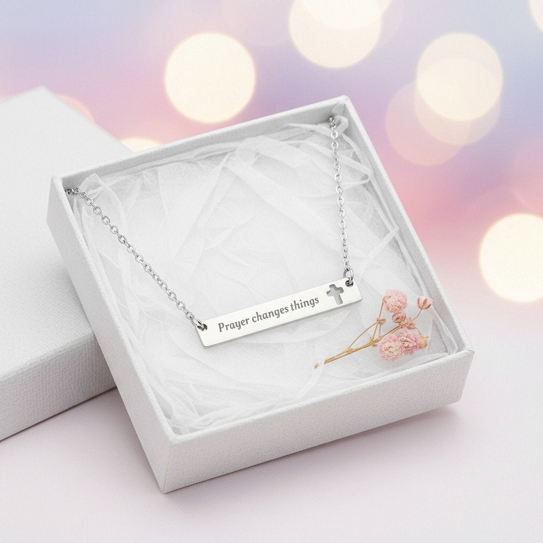 Silver engraved "Prayer Changes Things" bar necklace with cut-out cross displayed in open white gift box with white tissue and dried pink flowers, bokeh background, ready-to-gift Christian jewelry