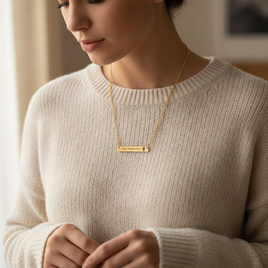 Woman wearing gold engraved "Prayer Changes Things" bar necklace with cut-out cross outdoors in garden, white blouse, soft sunlight, Christian jewelry lifestyle 