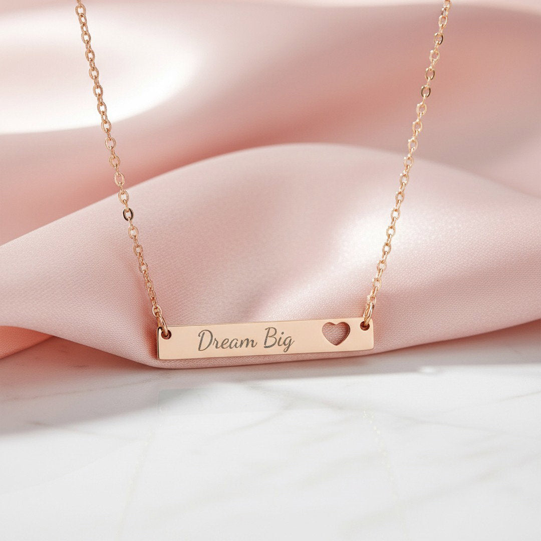 Rose gold Dream Big bar necklace with heart cutout displayed on soft pink satin fabric