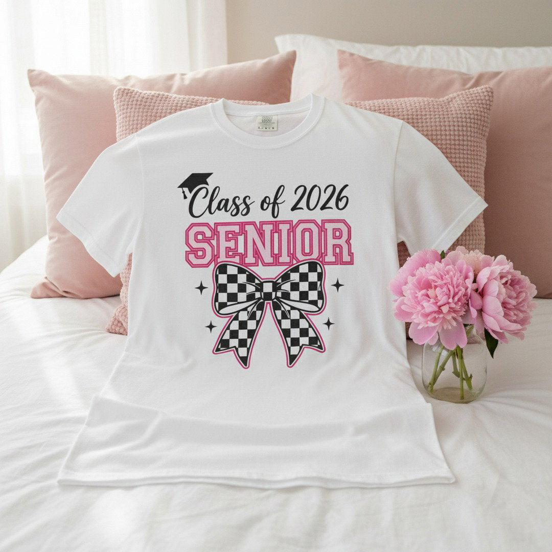 White Class of 2026 Senior Bow t-shirt laid flat on a white bed with pink throw pillows and pink peonies, bright airy lifestyle photo