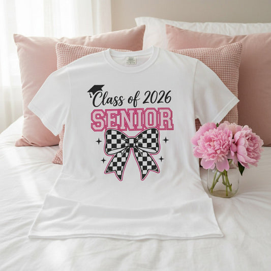 White Class of 2026 Senior Bow t-shirt laid flat on a white bed with pink throw pillows and pink peonies, bright airy lifestyle photo