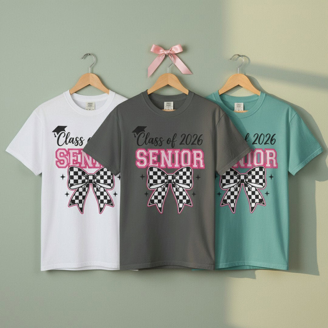 White, charcoal, and mint Class of 2026 Senior Bow graphic t-shirts hanging on wooden hangers against a sage green wall with pink ribbon accent