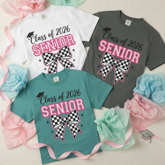 White, charcoal, and teal Class of 2026 Senior Bow t-shirts arranged in a flat lay with pastel pink, blue, and mint tissue paper flowers and a rhinestone graduation cap charm