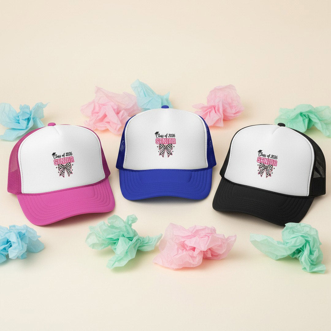 Three Class of 2026 Senior Bow trucker caps in pink, royal blue, and black arranged on cream background with pastel pink, blue, and mint tissue paper flowers, color options display