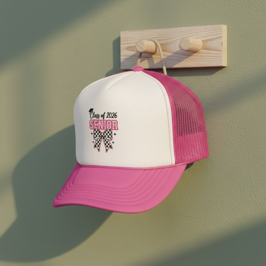 Pink and white Class of 2026 Senior Bow trucker cap hanging on a wooden peg rack against a sage green wall, soft natural light, graduation lifestyle photo