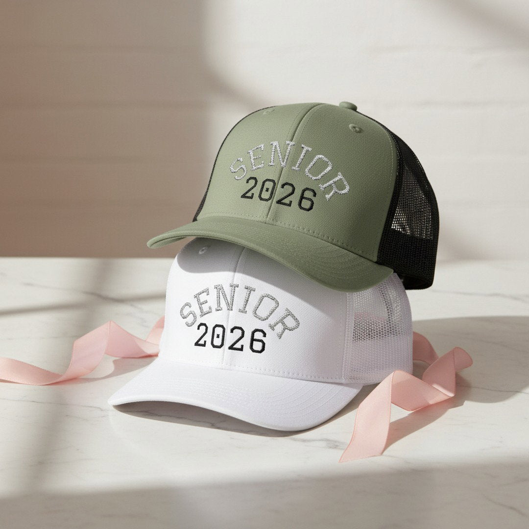 Two Senior 2026 embroidered trucker caps in sage green and white stacked on white marble surface with pink satin ribbon, graduation hat color options display