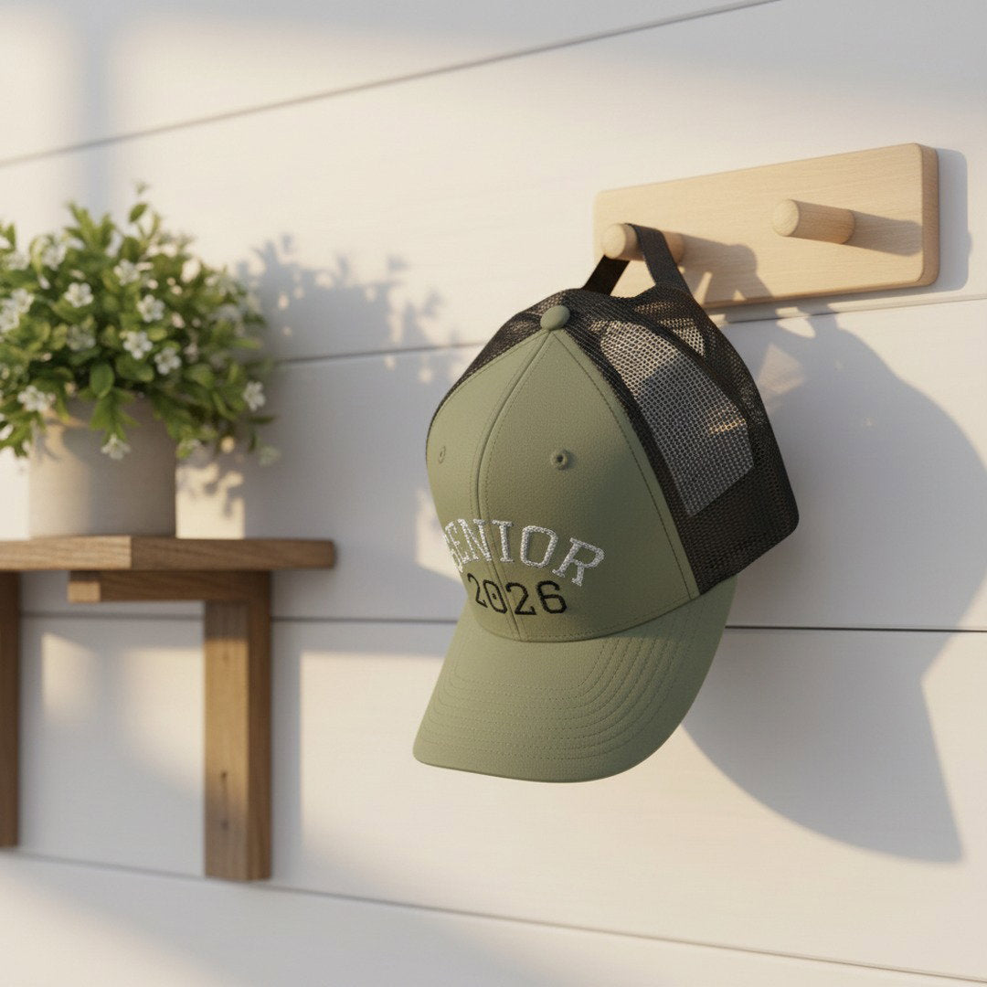 Sage green and black Senior 2026 embroidered trucker cap hanging on a wooden wall hook against a white shiplap wall with small white flowering plant beside it, graduation lifestyle photo