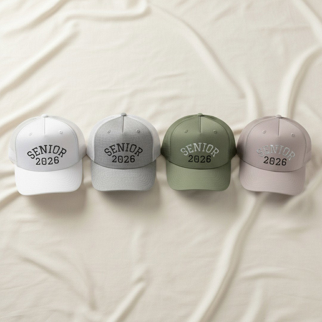 Four Senior 2026 embroidered trucker caps in white, heather grey, sage green, and light grey arranged in a row on cream linen, overhead flat lay showing all color options