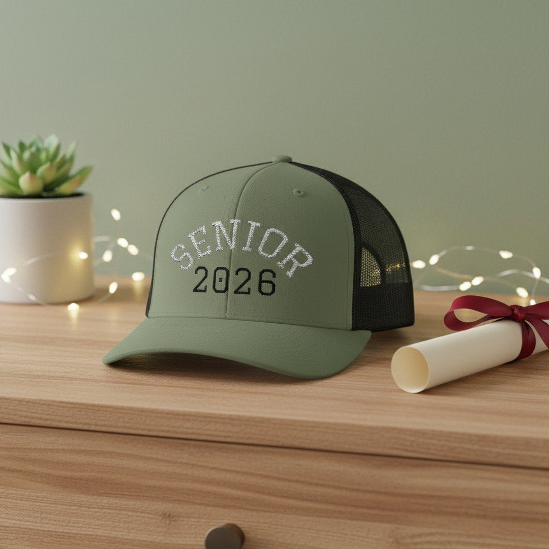 Sage green and black Senior 2026 embroidered trucker cap on a light wood dresser beside a rolled diploma with red ribbon and small succulent with fairy lights, cozy graduation gift display