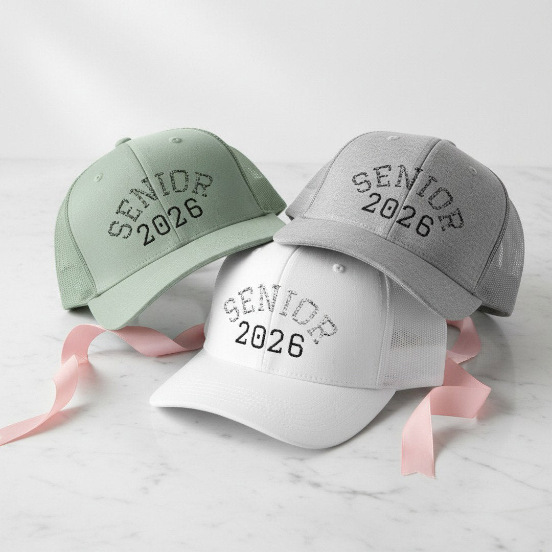 Three Senior 2026 embroidered trucker caps in sage green, heather grey, and white stacked on white marble surface with pink satin ribbon, graduation gift hat color options