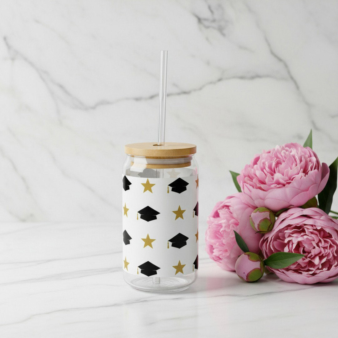 Graduation sipper glass with black grad caps and gold stars pattern on white marble surface beside pink peonies, bamboo lid and clear glass straw, feminine graduation gift idea